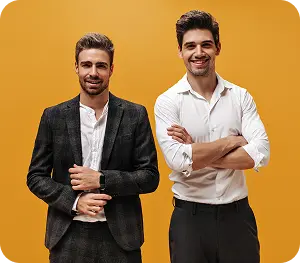 Sarı bir arka planın önünde, şık ve rahat iş kıyafetleri giymiş, kendinden emin iki erkek yan yana duruyor.